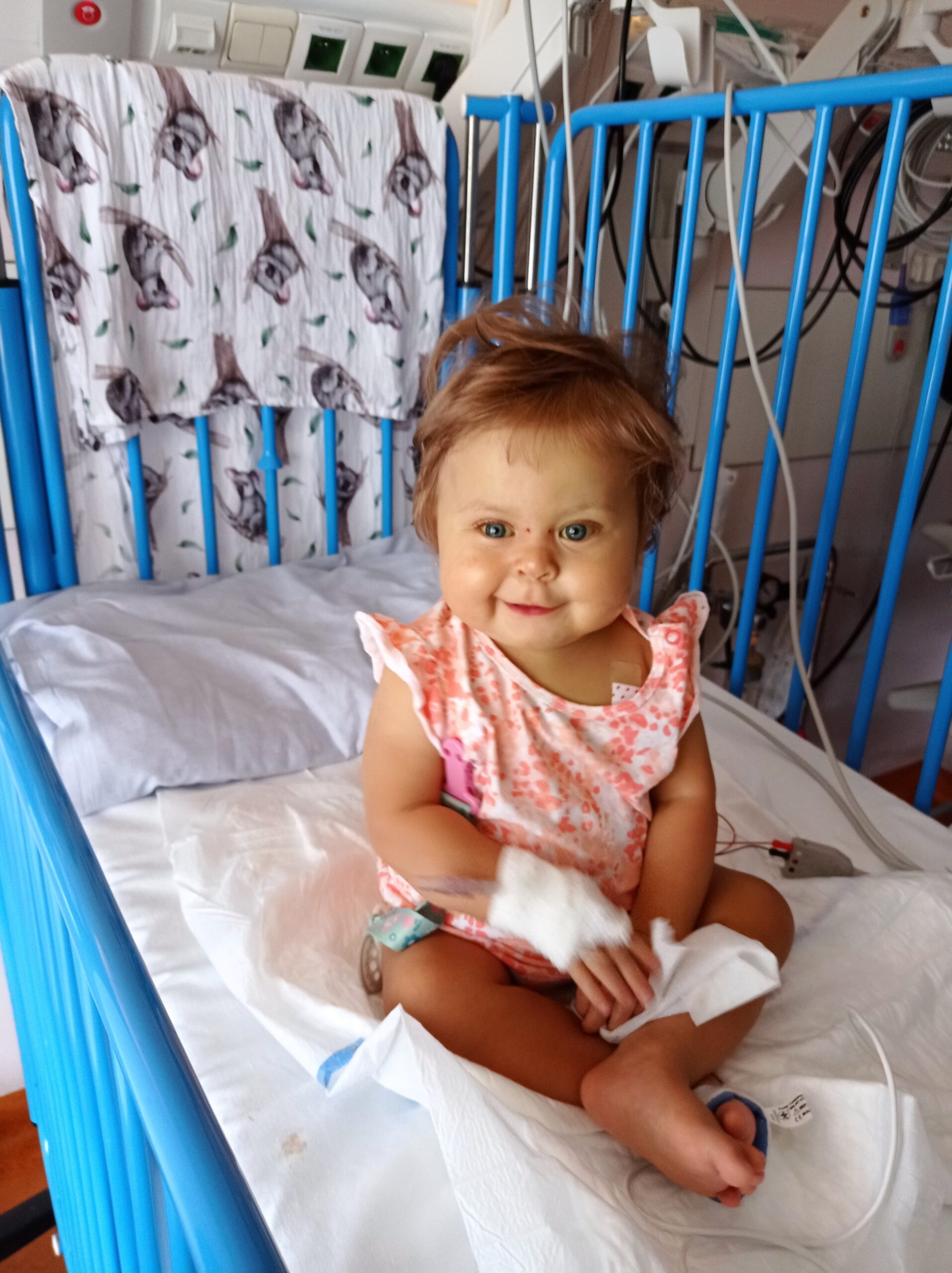 Mała dziewczynka Alicja Cielecka siedzi na łóżeczku szpitalnym i usmiecha się do zdjęcia mimo że podpięta jest do aparatury medycznej