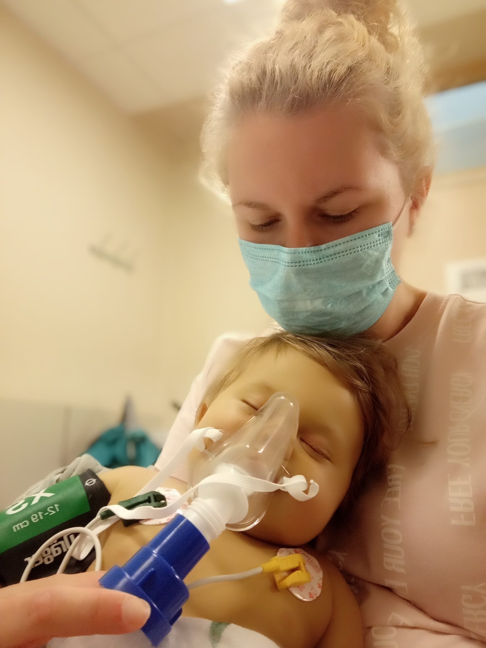 Mama z maseczką na twarzy trzyma małą dziewczynkę z przypiętymi elektrodami do ciałana rękach i przytrzymuje jej maskę tlenową